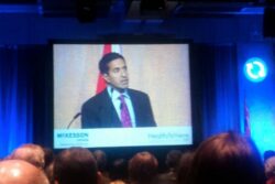 A man in a suit and tie stands at a podium, speaking at a conference. The backdrop includes a large screen displaying his image, with "McKesson Canada" and "HealthAchieve" logos visible. An audience is seated in front of the stage. lean blog