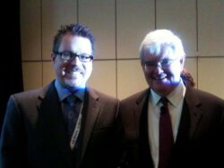 Two men stand side by side, smiling at the camera. Both are dressed in suits, with the man on the left wearing glasses and a blue tie, and the man on the right also wearing glasses and a darker suit with a red tie. The background is dimly lit and the image is slightly dark. lean blog