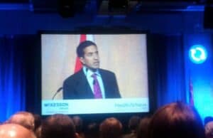 Some "Like Lean" Thoughts from CNN's Dr. Sanjay Gupta A man in a suit and tie stands at a podium, speaking at a conference. The backdrop includes a large screen displaying his image, with "McKesson Canada" and "HealthAchieve" logos visible. An audience is seated in front of the stage. lean blog