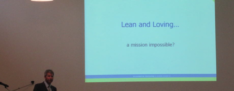A speaker stands at a podium addressing an audience, with a projected slide behind him titled "Lean and Loving... a mission impossible?" The slide has a green and blue border, and the setting appears to be a conference or seminar room. lean blog