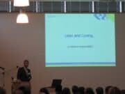 Lean is “Loving Care”? Lean IS Loving Care… Or Should Be A speaker stands at a podium addressing an audience, with a projected slide behind him titled "Lean and Loving... a mission impossible?" The slide has a green and blue border, and the setting appears to be a conference or seminar room. lean blog