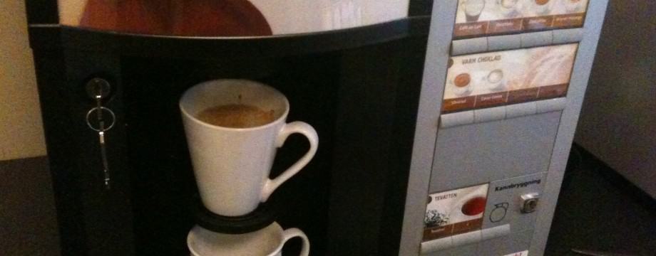 A coffee vending machine is dispensing coffee into two white mugs stacked on top of each other. The machine has a panel displaying options for different types of coffee beverages, and a small slot with a key on the left side. lean blog