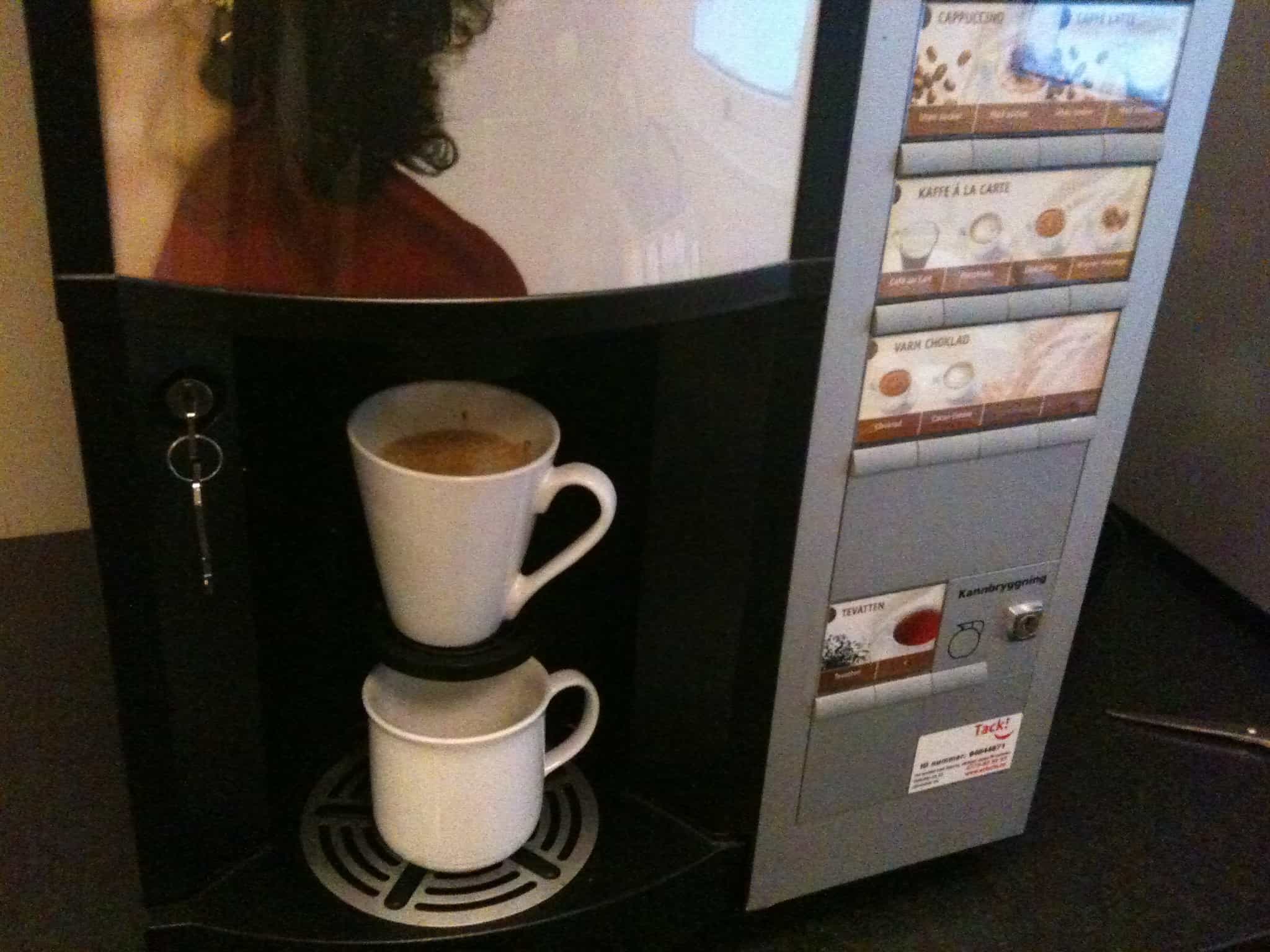 A coffee vending machine is dispensing coffee into two white mugs stacked on top of each other. The machine has a panel displaying options for different types of coffee beverages, and a small slot with a key on the left side. lean blog