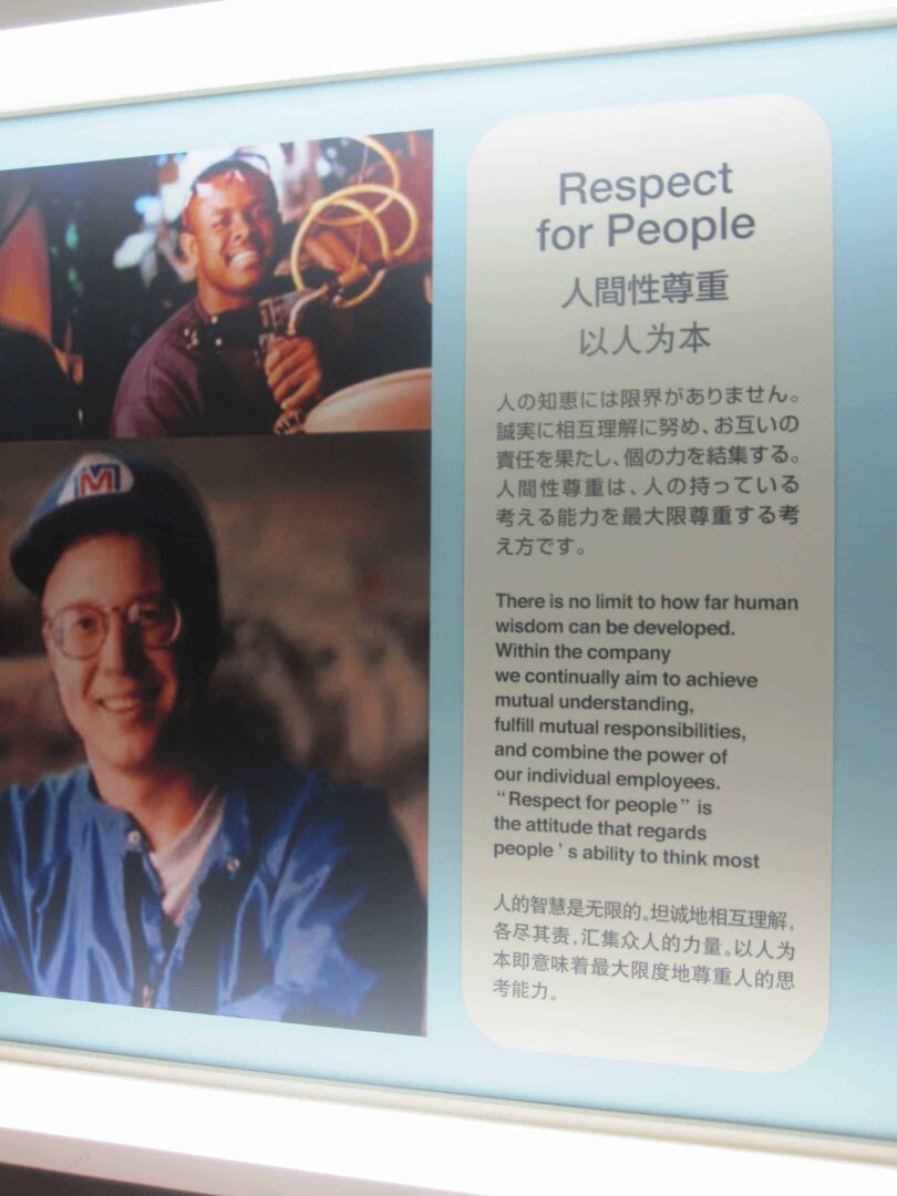 A Toyota-style display panel titled “Respect for People,” with Japanese and Chinese text below the heading. The right side explains the principle as developing human wisdom, pursuing mutual understanding, fulfilling mutual responsibilities, and combining the abilities of individual employees. On the left are two photos of employees, including one worker using a tool and another smiling worker wearing a blue uniform and cap. The display has a pale blue background and a clean museum or visitor-center style layout.