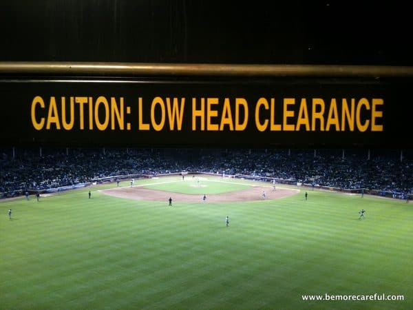Warning sign with yellow text CAUTION: LOW HEAD CLEARANCE centered over a night view of a professional baseball field at wrigley field chicago
