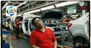 Prolonged worker resting at car assembly plant, with cars in progress and assembly workers in background.