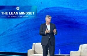 CEO Larry Culp’s Vision: Building a Lean Culture at General Electric A photo of GE Aerospace CEO Larry Culp standing on stage during a presentation. Behind him, there is a large screen with a blue background displaying the GE logo and the text "THE LEAN MINDSET: THE PURSUIT OF PROGRESS." Culp is wearing a dark suit and is gesturing with his hands while speaking. There is a chair and a table with a water bottle on the stage.