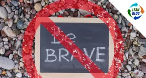 **Alt Text:** An image of a small chalkboard placed on a bed of multi-colored pebbles. The chalkboard has the words "Be Brave" written on it. Over the image, there is a red "No" symbol, a circle with a diagonal line through it, indicating a prohibition. In the top right corner, the Lean Blog logo is displayed, featuring a circular design with arrows in blue, green, and orange, and the text "LEAN BLOG" in the center.
