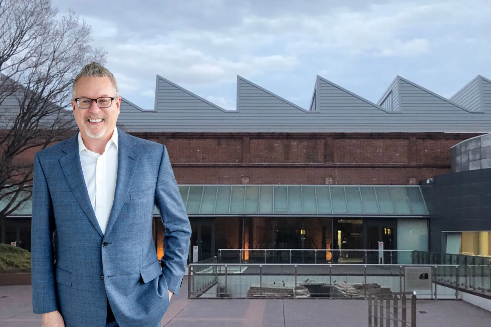 **Alt Text:** Mark Graban, wearing a blue suit and white shirt, smiling with his hands in his pockets, stands in front of the Toyota Commemorative Museum in Japan. The building features a distinctive zigzag roof design, large windows, and a glass entrance, with a courtyard and metal railing in front. The sky is overcast, and a leafless tree is visible on the left side of the image.