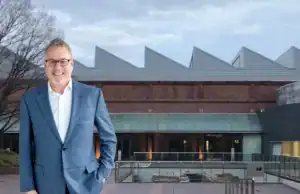 Come to Japan With Me and Katie Anderson in November to Study Lean! **Alt Text:** Mark Graban, wearing a blue suit and white shirt, smiling with his hands in his pockets, stands in front of the Toyota Commemorative Museum in Japan. The building features a distinctive zigzag roof design, large windows, and a glass entrance, with a courtyard and metal railing in front. The sky is overcast, and a leafless tree is visible on the left side of the image.