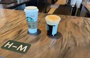Why Starbucks’ Inconsistent Mobile Order Process Frustrates Customers **Alt Text:** A Starbucks counter displaying two drinks labeled "Mark." On the left is a tall hot coffee in a white cup with a sleeve, and on the right is a Nitro Cold Brew in a clear plastic cup with a domed lid, filled with a frothy cream top. The drinks are placed on a dark, textured countertop with a section labeled "H-M" in white letters. The background shows a part of the store's interior with coffee equipment and a barista in the distance.