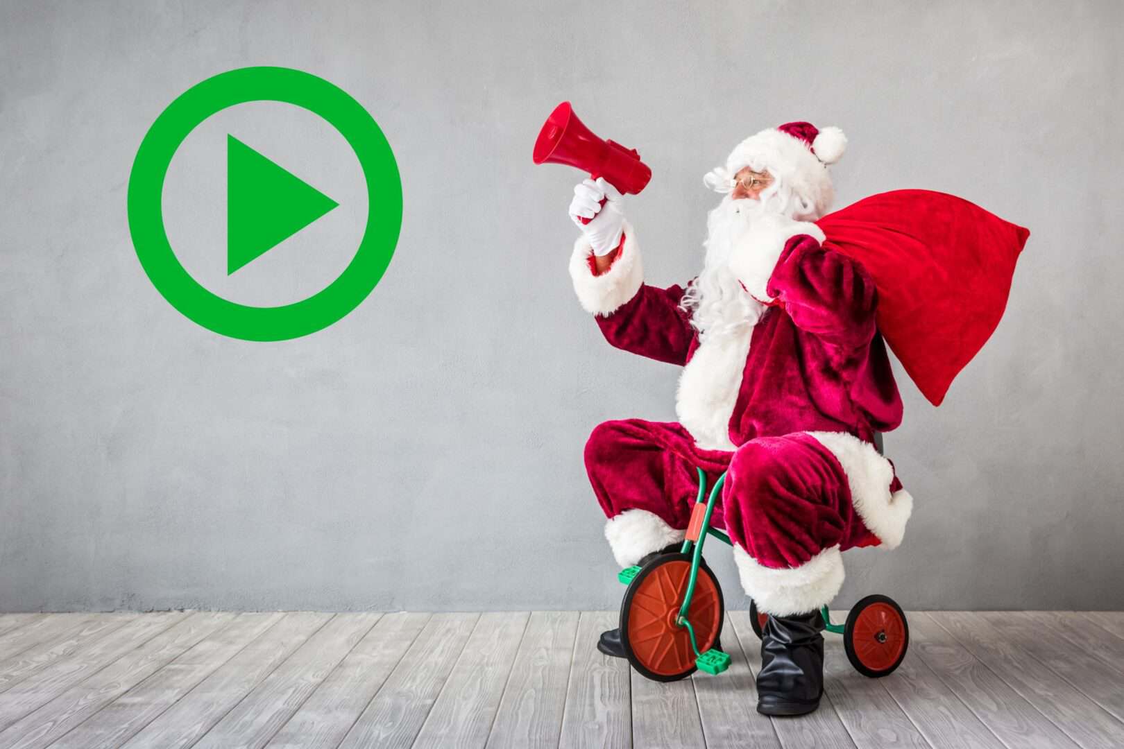 Play button icon next to Santa riding a tricycle with a large red sack, against a plain wall background.