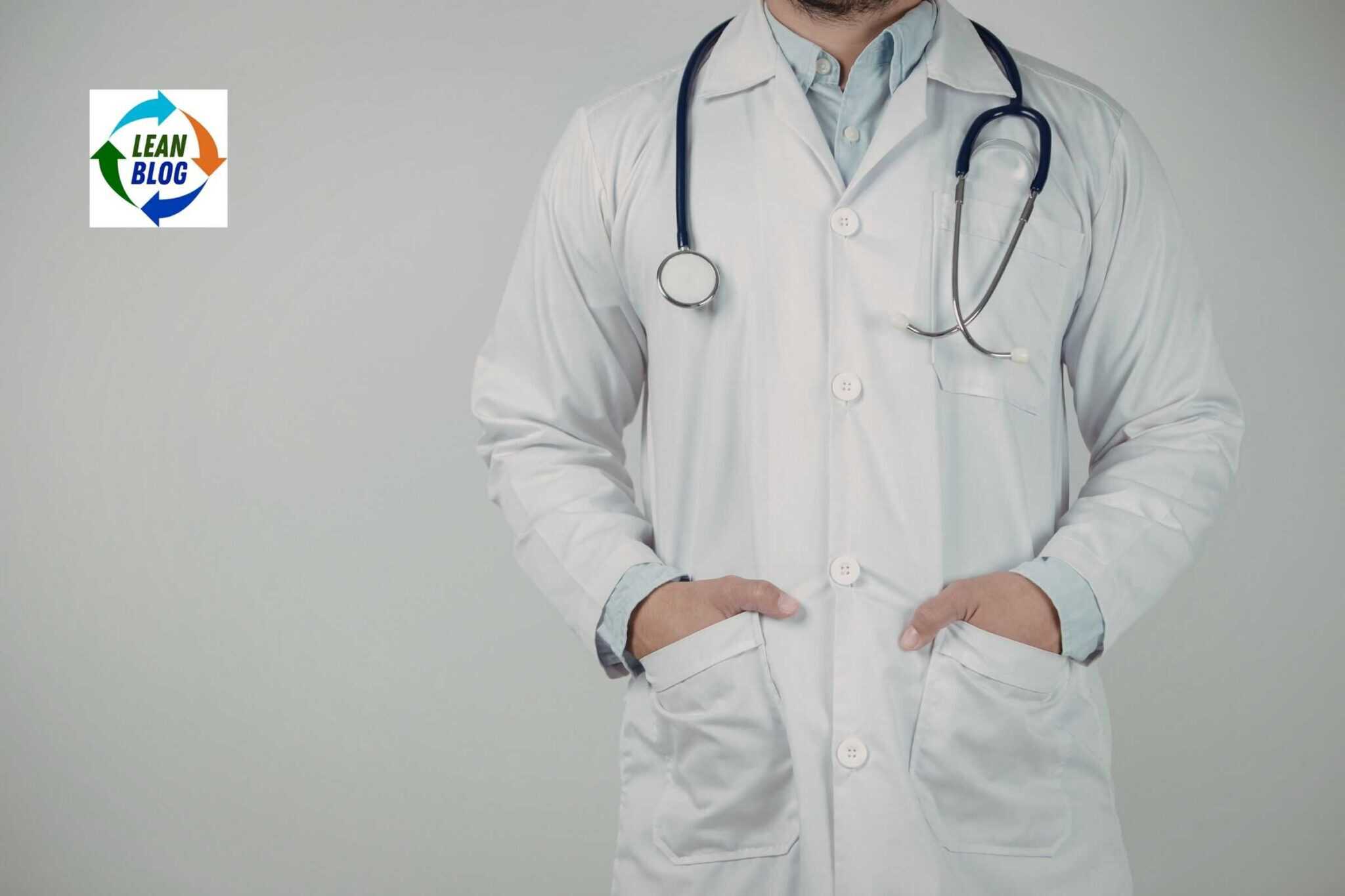 Doctor in a white coat with stethoscope, hands in pockets, against plain background.
