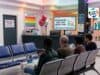 The ER Wait Time Metric That Can Hide Deteriorating Patient Experience A bright hospital emergency room waiting area where the space is decorated with red and silver balloons and colorful lighting. Four patients are seated in blue chairs, with one reading a pamphlet. A large TV screen behind the reception counter reads "CELEBRATING WAIT TIME REDUCTIONS! Our Shortest Wait Ever!" accompanied by a bar graph and colorful confetti. Various posters on the walls list the "TRIAGE PROCESS & EXPECTATIONS" and celebrate achievements like "NEW RECORD: TRIAGE IN < 10 MINUTES" and "45% AVERAGE WAIT REDUCTION."