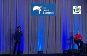 The Most Misunderstood Role in the Toyota Production System Michelle Thomas and Jamie Bonini of Toyota's Production System Support Center (TSSC) speaking on stage at the 2026 Lean Summit, with blue uplighting and a gray curtain backdrop.