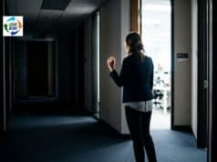 What Can a CI Director Do When Executives Undermine Psychological Safety? Person hesitating in a corporate hallway outside an open office door with bright fluorescent light spilling through the doorway.