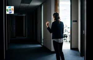 What Can a CI Director Do When Executives Undermine Psychological Safety? Person hesitating in a corporate hallway outside an open office door with bright fluorescent light spilling through the doorway.