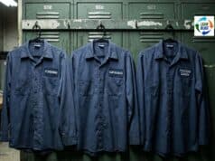 Ford Pays Process Coaches Six Figures. They Quit Within Eighteen Months. A close-up of a metal name badge or nameplate on a dark blue work shirt. The badge reads "PROCESS COACH" in crisp embossed letters. The shirt and badge look new. Behind the figure, slightly out of focus, is an industrial factory environment with fluorescent overhead lighting and assembly line equipment. The composition emphasizes the contrast between the pristine, almost promotional quality of the title on the badge and the harsh, utilitarian reality of the environment. Photorealistic, documentary style, muted industrial color palette with deep blues and steel grays. The mood is quietly ironic - the title and the setting don't match.