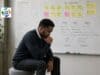 What Do You Know? And How Do You Know It? Man seated in Rodin Thinker pose in front of a whiteboard covered with A3 problem solving, fishbone diagram, and sticky notes.