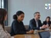 Four Recent Episodes on Why People Stay Silent — and What It Costs A quiet office meeting in a modern conference room, with one woman in sharp focus looking down with folded hands while colleagues around her continue the discussion in soft blur.