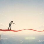 Manga- and anime-influenced editorial illustration in a wide horizontal format. A small lone figure, seen from behind in plain clothes, stands on a glowing crimson ribbon that stretches horizontally across the frame like a floating path. The ribbon gently rises and falls but stays level. The person leans forward with one arm extended, reaching toward a soft sage-green glowing ring far below and ahead across a wide empty gap. Pale cliffs frame the distant lower edges, while most of the image is filled with a vast cream-to-blue sky with thin clouds and ample negative space. The mood is quiet, reflective, and slightly wistful.