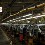 Alt text: A yellow pull cord hangs untouched above an automotive assembly line while workers continue building vehicles in the background.