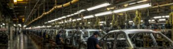 Alt text: A yellow pull cord hangs untouched above an automotive assembly line while workers continue building vehicles in the background.