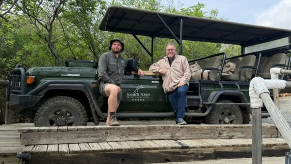 Two men sitting on the side of a green open-air safari vehicle with Namibti Plains Private Game Reserve branding, parked on a wooden deck.