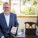 Mark Graban in a blue suit holding a tablet displaying the book cover The Mistakes That Make Us