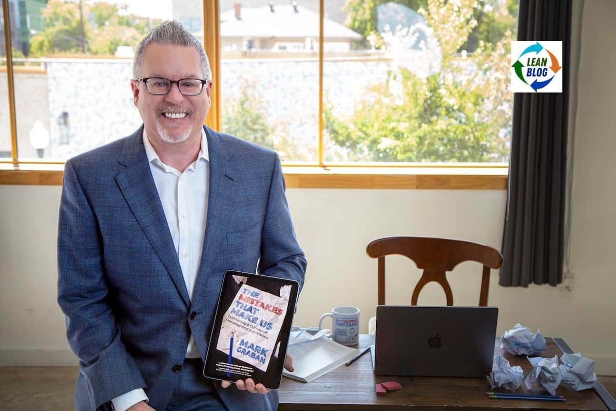 Mark Graban in a blue suit holding a tablet displaying the book cover The Mistakes That Make Us