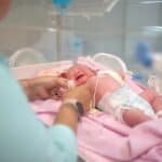 Person in scrubs adjusting medical leads on a crying infant lying on a pink blanket inside a hospital incubator