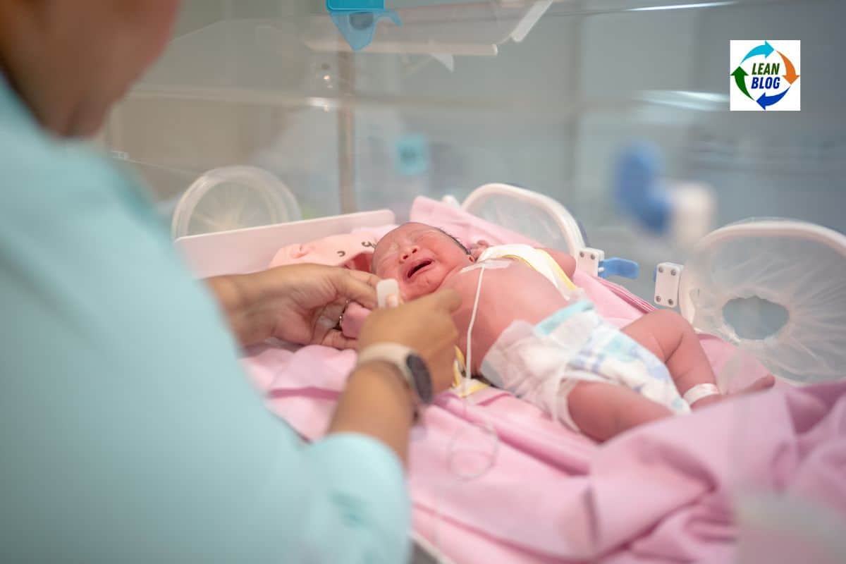 Person in scrubs adjusting medical leads on a crying infant lying on a pink blanket inside a hospital incubator