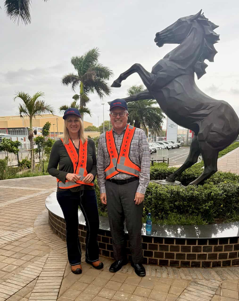 Alt text: Melanie Veness and Mark Graban in orange VISITOR vests and Toyota caps beside a horse statue outside Toyota's Durban plant